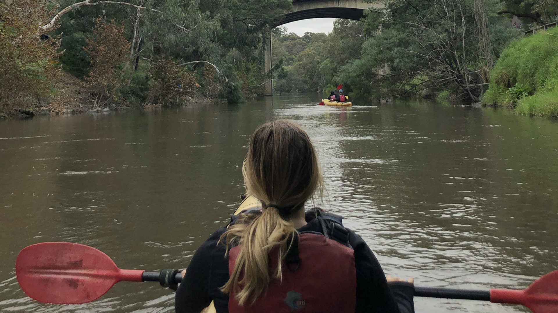 A person with long hair in a ponytail floats in a kayak in the middle of a river. There is a bridge in the distance at the top of the image and trees either side
