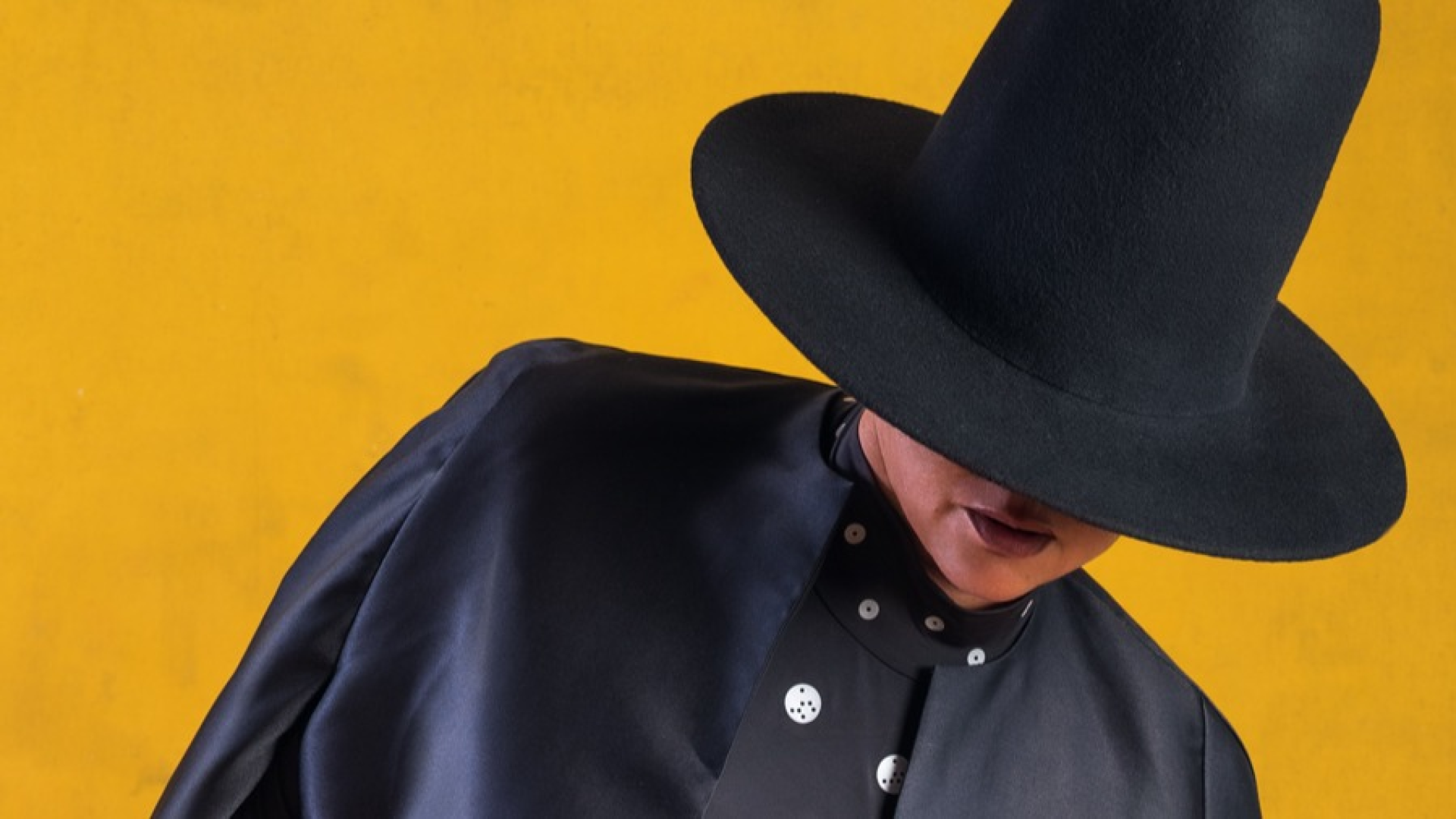 Person in high crown black hat, black cape against a yellow background