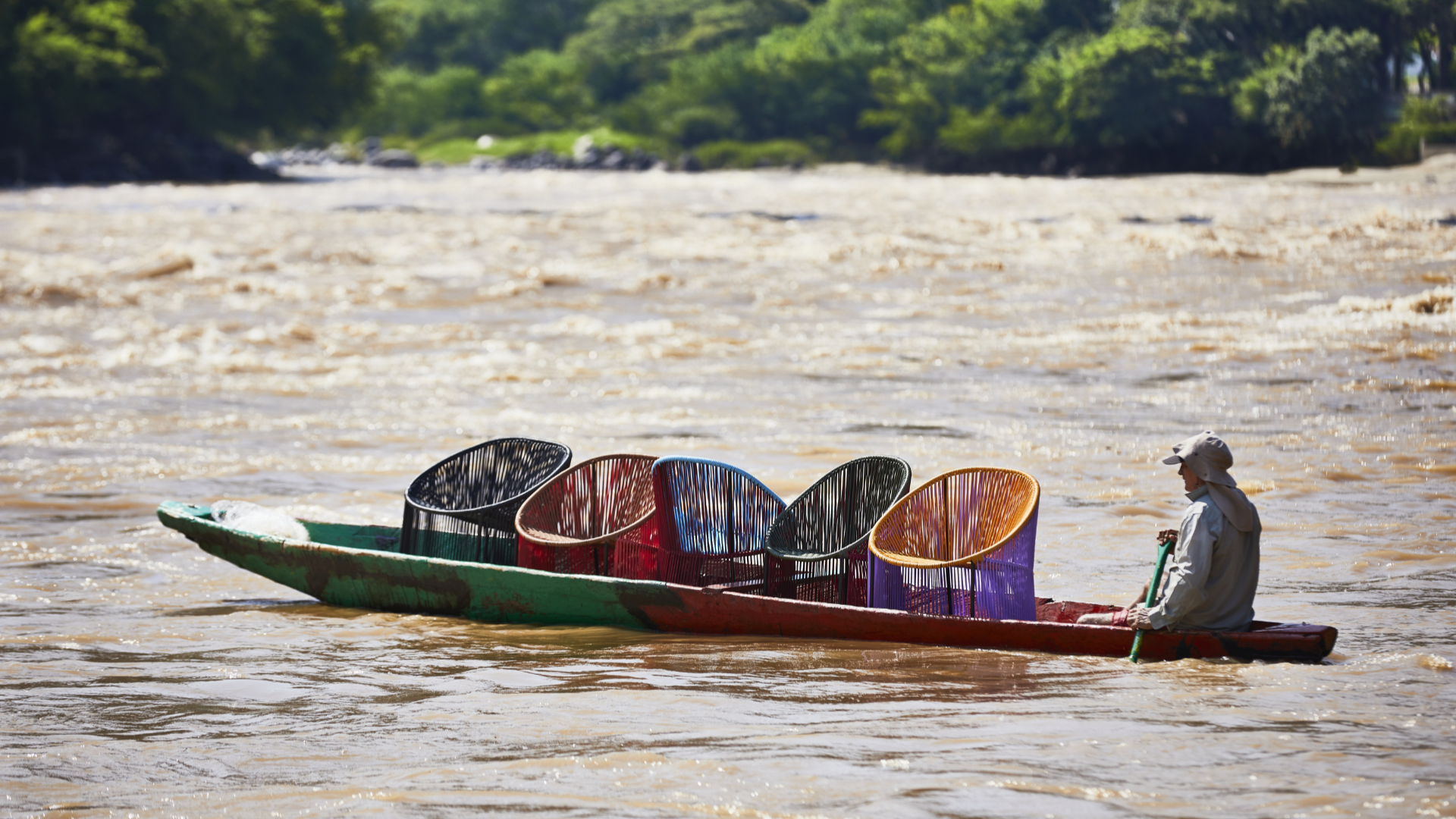 Image of five lounge chairs being transported across a river in Columbia by canoe. 
