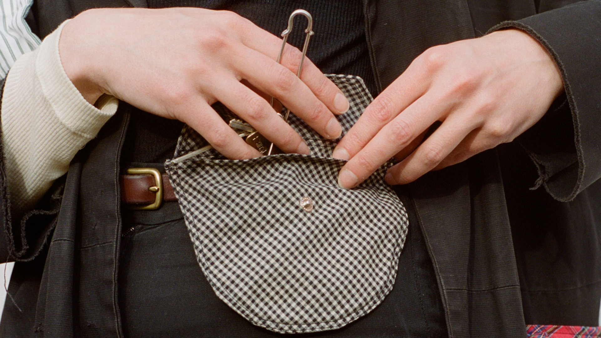 A close up of a women wearing a black and white check Future Archive attachable pocket bag on a brown leather belt over a black trousers and a black and white top. One hand is holding the pocket flap open and the other reaching in holding keys.