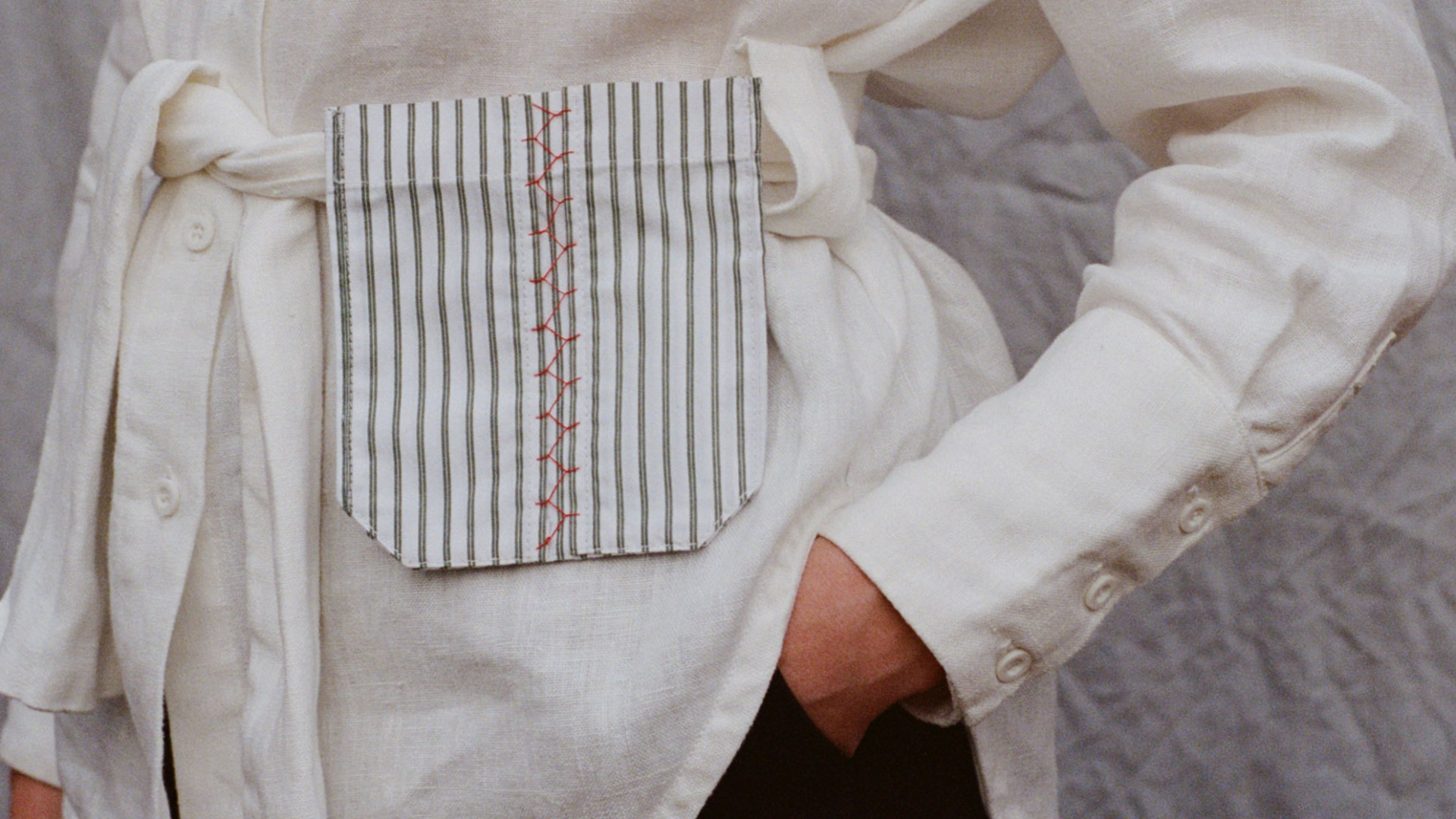 A close up of a women standing wearing a green and white striped Future Archive attachable pocket bag with red decorative stitch detailing on the waist tie of a white linen shirt. One hand is entering her pocket trousers and the other sits relaxed at her side. 