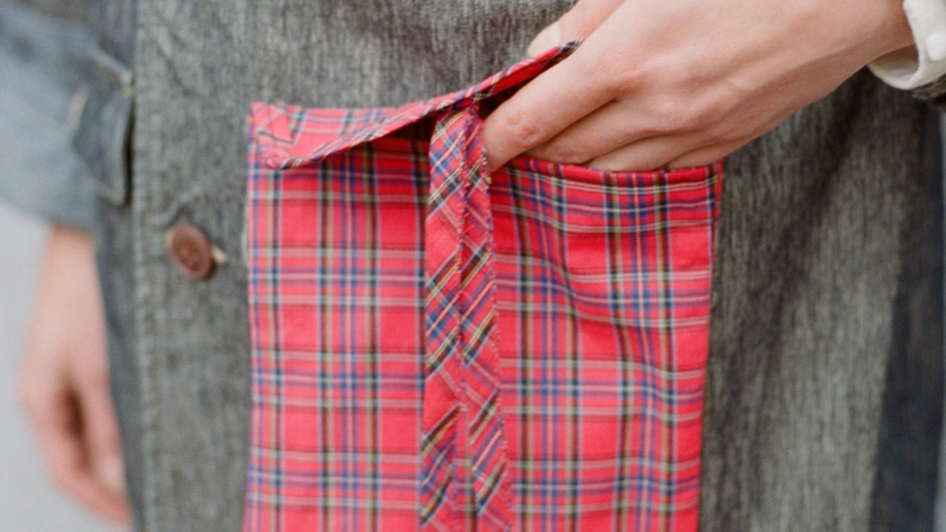 A close up of a women standing wearing a red tartan Future Archive attachable pocket bag on a denim coat with brown buttons. One hand is entering the attachable pocket and the other is relaxed at her side.
